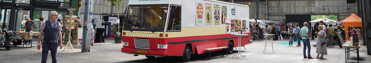 SRV-wagen staat midden in beeld, in een grote loods of hal, eromheen en op de achtergrond staan mensen en kraampjes van het festival