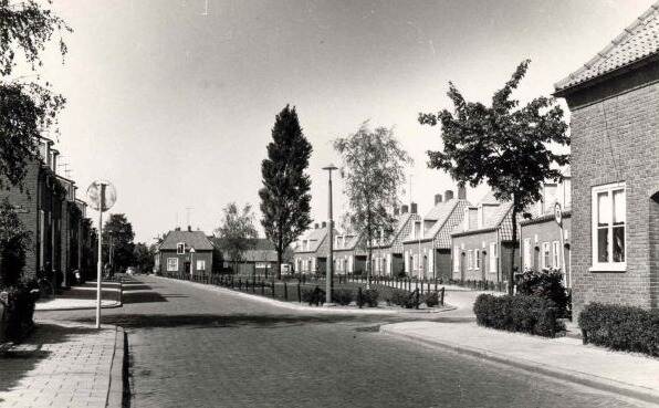 Historische zwart-wit foto van een straat, in het midden een klein plantsoen met daaromheen huizen