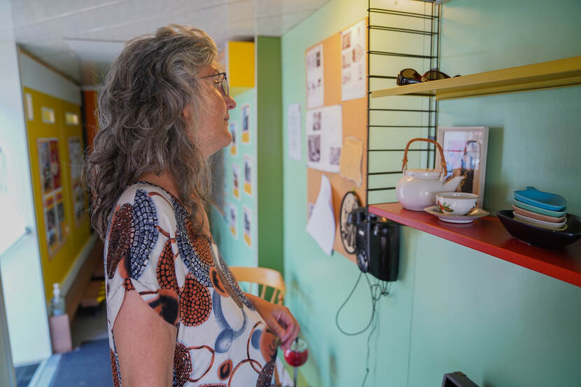 Een mevrouw bekijkt een wandrek met daarop o.a. een theeservies, aan de muur hangt een oudedraaitelefoon