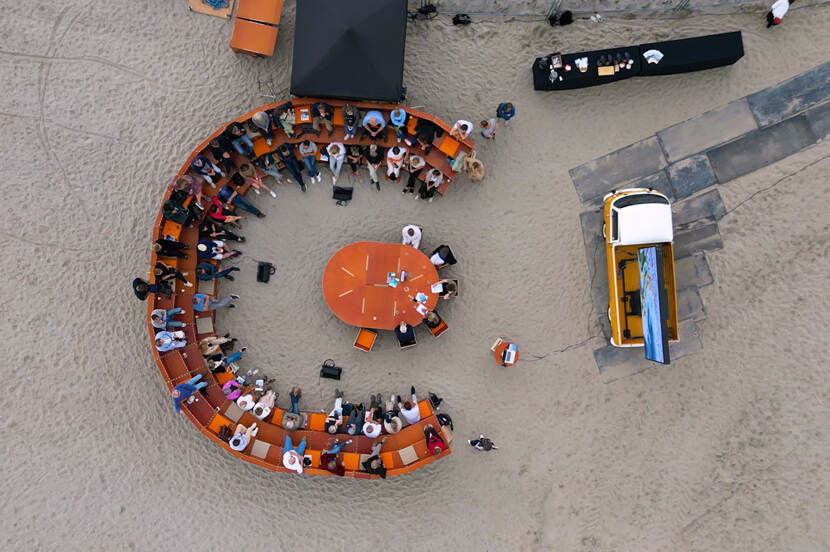 Luchtfoto van een c-vormige tribune op het strand van Hoorn met mensen erop. De tribune staat rondom een tafel waar een gesprek plaatsvindt.