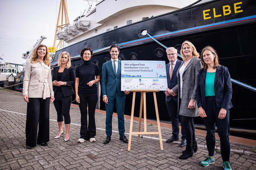 De partners van de Erfgoed Deal, inclusief de minister en staatssecretaris, staan naast een bord waarop het ministerie van Economische Zaken en Klimaat en de Unie van Waterschappen net een handtekening gezet hebben. Het gezelschap staat voor een boot in de haven van Maassluis.